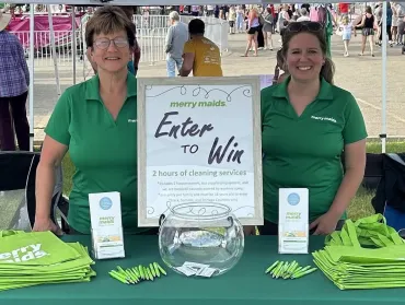 two smiling women in green merry maids shirts standing by an enter to win sign at a fair booth